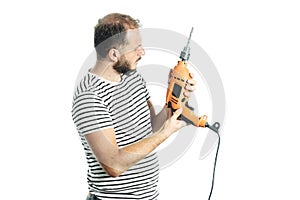 A bearded handyman in a striped T-shirt looking at the drill machine in his hand with a questioning look.