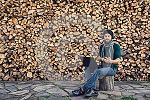 Bearded hipster freelancer in headphones working on laptop and uses phone against background of firewood wall