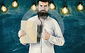 Bearded hipster holds book, chalkboard on background. Recommendation concept. Teacher in eyeglasses recommends book