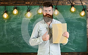 Bearded hipster holds book, chalkboard on background. Teacher in eyeglasses recommends book, thumbs up. Recommendation