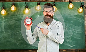 Bearded hipster holds clock, chalkboard on background. . Teacher in eyeglasses holds alarm clock and pen. Discipline