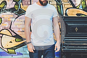 Bearded hipster man dressed in white t-shirt is stands against wall with graffiti. Mock up. Space for logo, text, image.