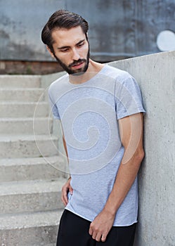 A bearded hipster man in a gray T-shirt, pants pants posing against a gray stone wall in the afternoon.