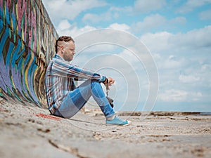 Bearded hipster man with mohawk sits on a concrete pier near the sea and looks into the distance against the sky and clouds