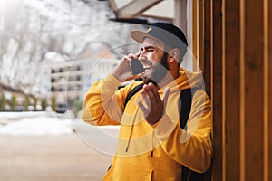 Bearded hipster man tourist in yellow hoodie and cap stands outdoors, talking on mobile phone. Smiling man calling friends