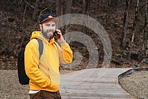 Bearded hipster man tourist in yellow hoodie and cap stands outdoors, talking on mobile phone. Smiling man calling friends