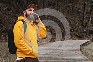 Bearded hipster man tourist in yellow hoodie and cap stands outdoors, talking on mobile phone. Smiling man calling friends