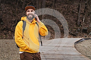 Bearded hipster man tourist in yellow hoodie and cap stands outdoors, talking on mobile phone. Smiling man calling friends