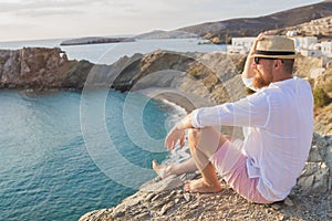 A bearded hipster man on vacation sits on a steep seashore and looks into the distance