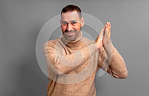 Bearded Hispanic man wearing a beige turtleneck clapping his hands proud and pleased at the spectacle he just witnessed
