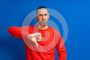 Bearded hispanic man wearing red sweater sulking shows thumb down with negative attitude, isolated over blue background.