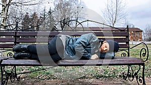 Bearded homeless man lies on a bench to sleep in a city park. Below poverty line. Male tramp in dirty clothes with cap and hat.