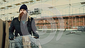 Bearded homeless man walking down the street with shooping card at cold autumn day