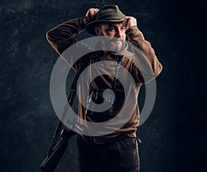 Bearded hunter with rifle and binoculars corrects his hat and looking sideways. Studio photo against a dark wall