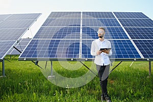 Bearded male engineer using tablet to control photovoltaic panels remotely during work on solar farm on sunny day