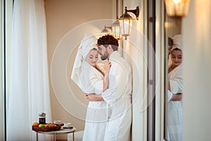 Bearded male and his wife wears white bathrobes and towel on head, hug each other, feel relaxed after taking bath in