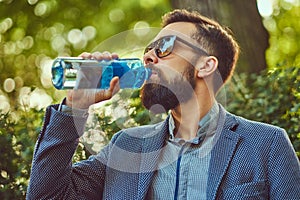 Bearded male man drinking cool water outdoors, sitting on a bench in a city park.