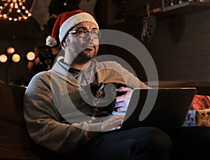 Bearded male in Santa`s hat dreaming on a couch with a laptop.