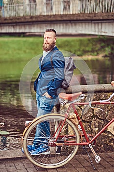 Bearded male with a stylish haircut dressed in casual clothes with a backpack, standing with a retro bicycle near the