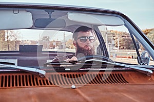 Bearded male in sunglasses dressed in brown leather jacket driving a retro car.