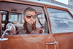 Bearded male in sunglasses dressed in brown leather jacket driving a retro car.