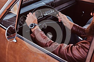 Bearded male in sunglasses dressed in brown leather jacket driving a retro car.