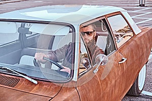 Bearded male in sunglasses dressed in brown leather jacket driving a retro car.