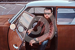 Bearded male in sunglasses dressed in brown leather jacket sits behind the wheel of a tuned retro car with open door.