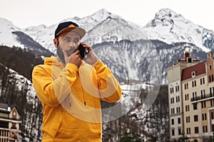 Bearded male tourist in yellow hoodie and cap stands on background of high snowy mountains, talking on mobile phone. Lifestyle