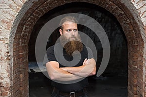 A bearded man with arms crossed, stands in a gloomy room under a curved arch