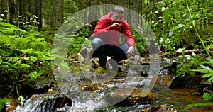 A bearded man with a backpack drinks water from a Mountain stream