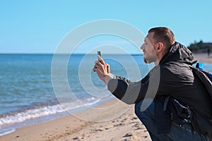 Bearded man in black raincoat on a blue sea landscape background with smartphone in hands takes selfie photography