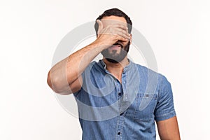 Bearded man in blue shirt closing eyes with hand, dont want to see that, ignoring problems, hiding from stressful situations