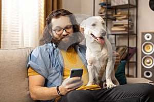A bearded man browses the internet on a smartphone in the company of his dog. A contented middle-aged boy uses his phone