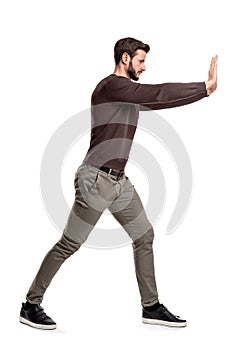 A bearded man in casual clothes tries to push a heavy object with both arms with one leg put in front for balance.