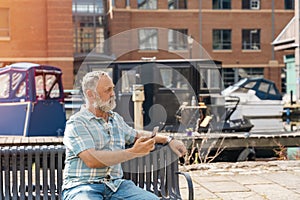 bearded man chatting by phone on the quays, pier, sitting on bench, looking at yacht, sailboat, boat. Lifestyle concept