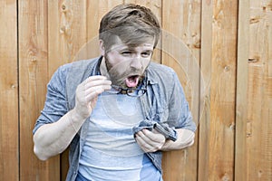 Bearded man cleans his glasses from dust and is going to wipe them with a shirt.  on the background of an old wooden wall