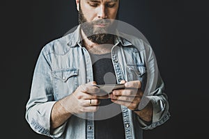 Bearded man in denim shirt holding smartphone in hands and texting. Young hipster using mobile phone for online communication