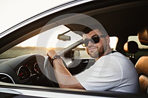 Bearded man driver in sunglasses sitting in his car and looking at camera