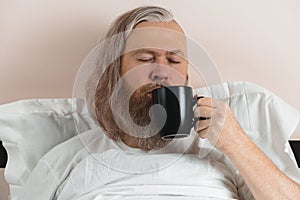 Bearded man enjoying his morning coffee in bed to cheer up before starting new day. Closeup