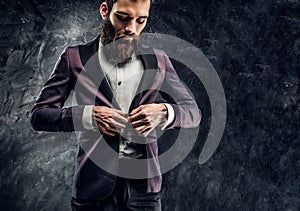 A young bearded man with expressive style buttoning his elegant jacket standing against a dark textured wall