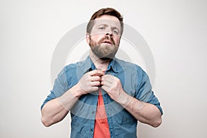 Bearded man fastens buttons on his shirt and looks haughtily at the camera, on a white background photo