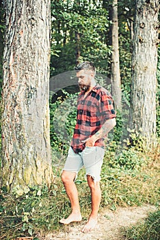 Bearded man in forest. Hipster with long beard on natural green landscape. Tourist in plaid shirt and jean shorts travel