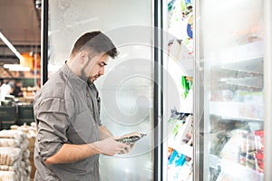 Bearded man with frozen food in his hands stands at the refrigerator in the department of frozen food. Buyer selects and purchases