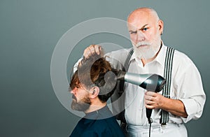 Bearded man getting groomed by hairdresser with hair dryer at barbershop. Serios hairdresser holding a blow dryer.