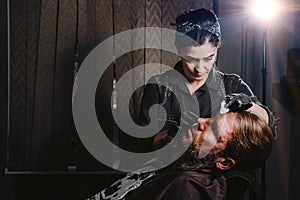 Bearded man getting haircut by hairdresser with dangerous razor at barbershop