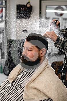 Bearded man getting hairstyle. Barber gently splashes water from the spray bottle at the head hair of client.