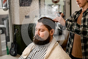 Bearded man getting hairstyle. Barber splashes water from the spray bottle at the head hair of client.