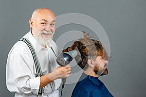 Bearded man getting hairstyle by hairdresser with hair dryer at barbershop. Funny hairdresser holding a blow dryer.