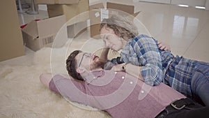 Bearded man in glasses and pretty woman with curly hair lying together on fluffy carpet and talking close up. Unpacked